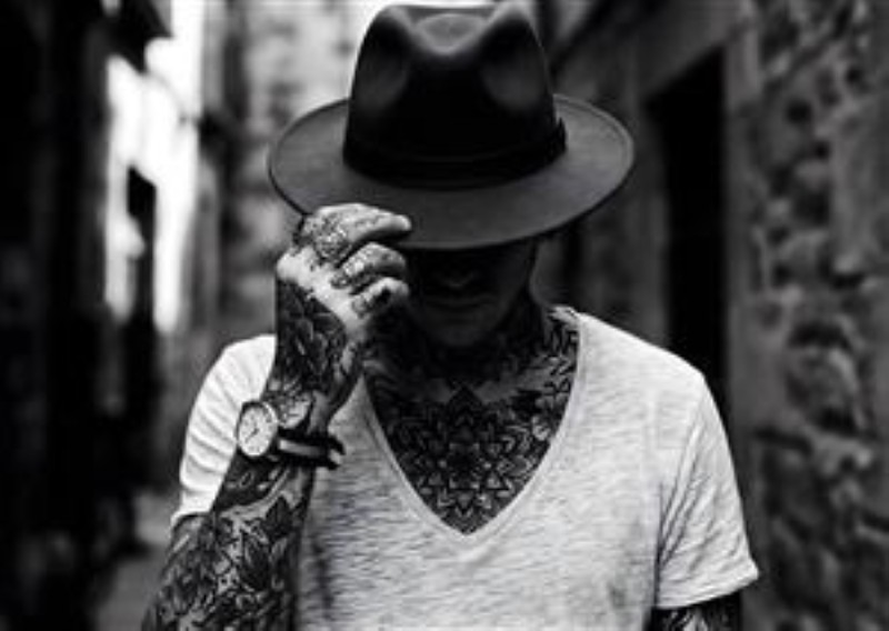 Tatoveringer og Stilfuld Hat - Oplev det rå og kunstneriske udtryk med dette nærbillede af en person, der bærer en elegant hat og imponerende tatoveringer. Det sløret baggrund tilføjer dybde og fokus på den unikke kropskunst og den afslappede stil.