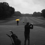 Golfspiller svinger en golfkølle med en golfvogn i baggrunden og en person med en gul paraply.