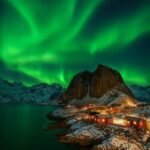 Nordlys i grønne nuancer over en fjord med røde hytter og bjerge i baggrunden.
