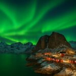 Nordlys oplyser nattehimlen over røde hytter ved en sø i bjerglandskab.