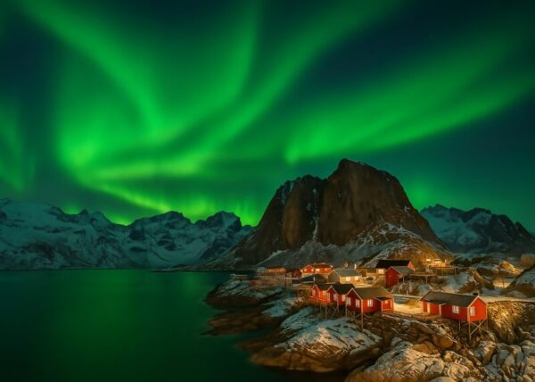 Nordlys oplyser nattehimlen over røde hytter ved en sø i bjerglandskab.